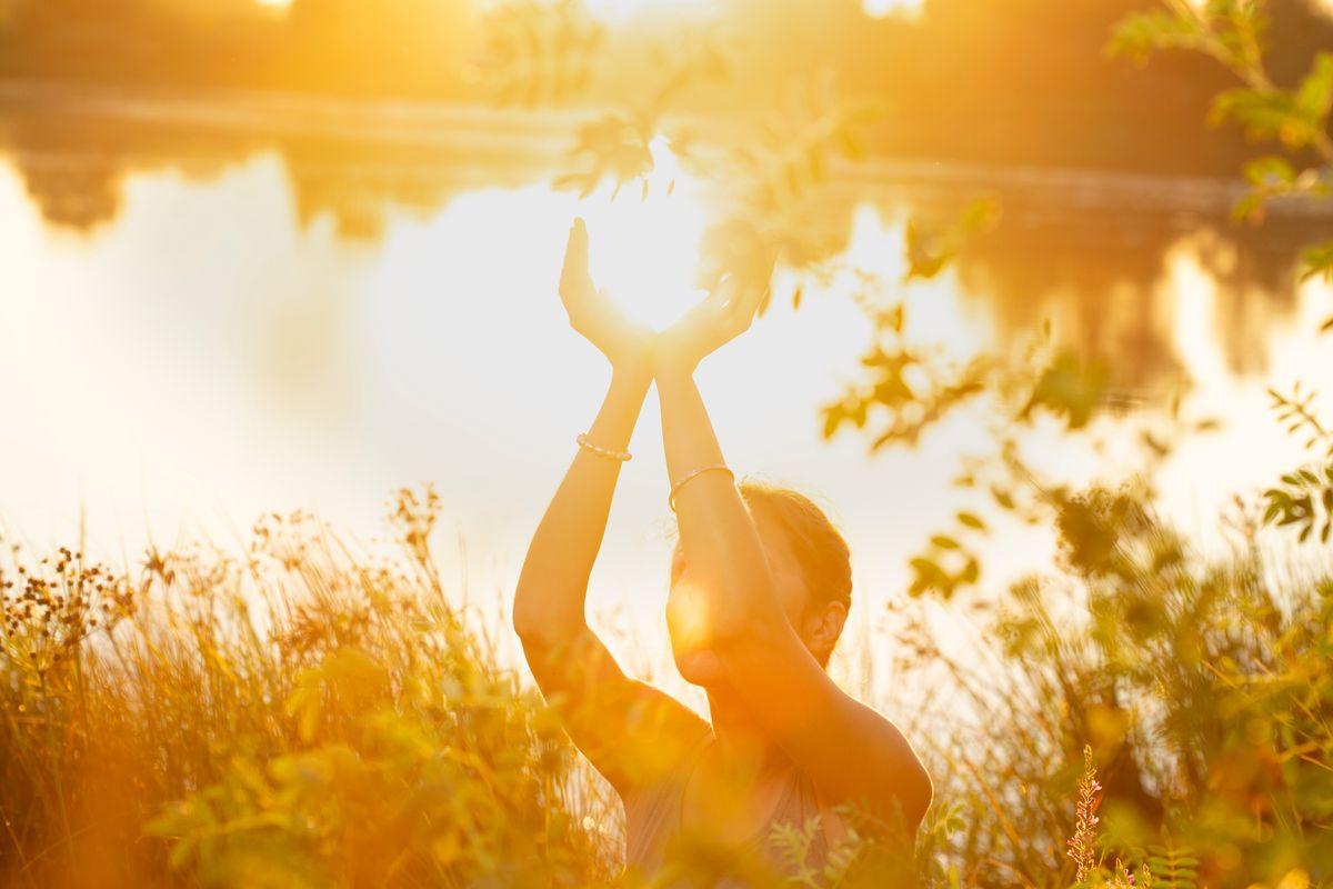 Woman yoga hands in prayer position and  heart shaped palms, blooming spring lush meadow with green grass in lighting of sunset  on the lake.